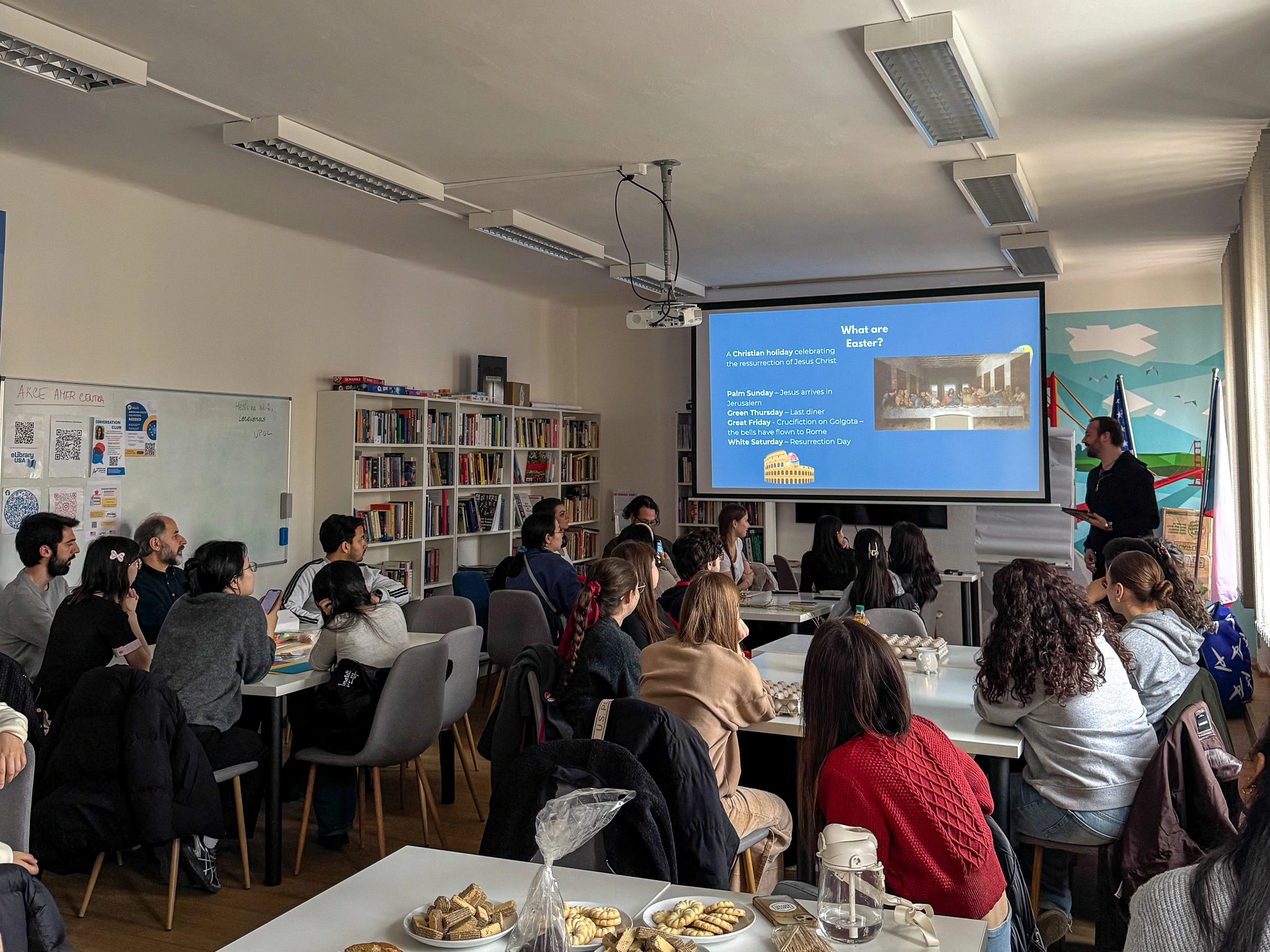 A room with diverse university students seated around tables, attentively listening to a presenter beside a screen displaying "What are Easter?" The atmosphere is focused and academic.